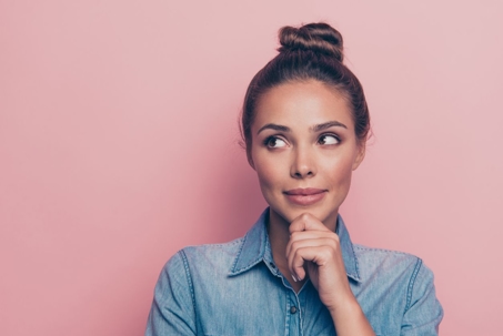 A woman with her hair in a bun holds her chin in her hand, thinking. She is wearing a blue jean shirt and is standing in front of a pink wall.