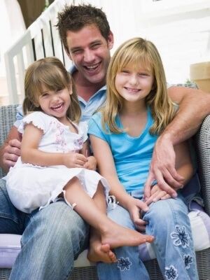 A father with visitation rights sits on stairs with his two young daughters on his lap. All three are smiling at the camera.