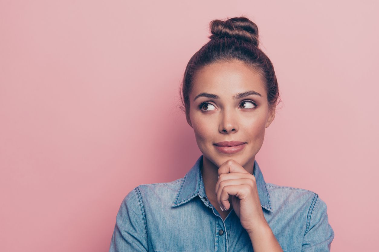 A woman with her hair in a bun holds her chin in her hand, thinking. She is wearing a blue jean shirt and is standing in front of a pink wall.