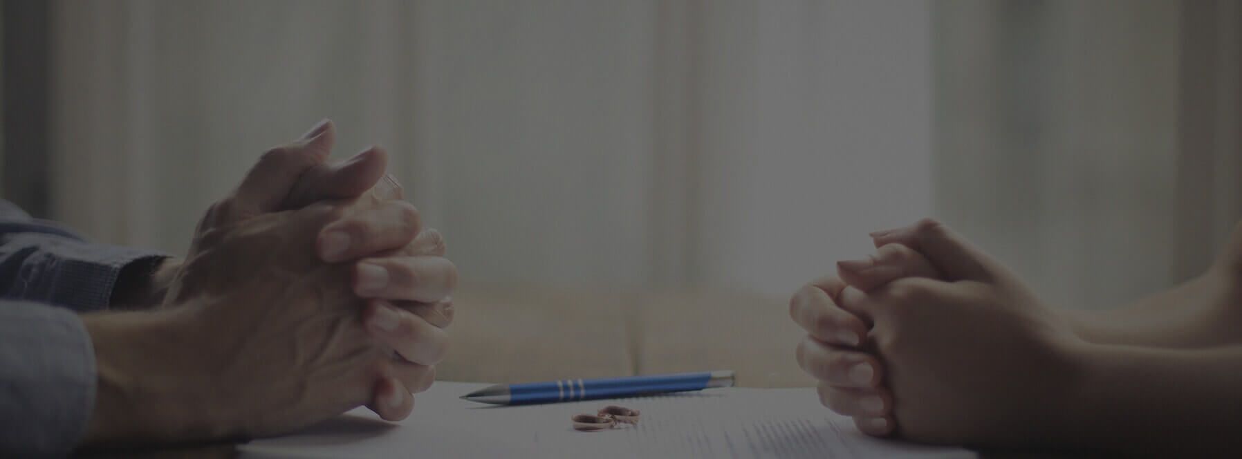 Two pairs of folded hands rest on a table across from each other. There are divorce papers, two golden wedding bands, and a pen laying on the table between the two people.
