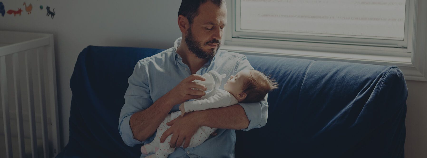 A father sits on a blue couch feeding his newborn child